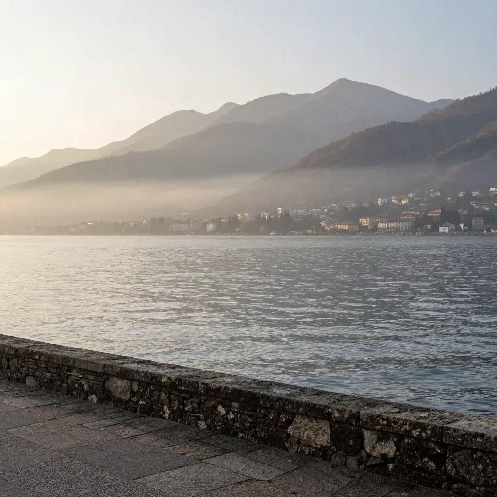 Lago di Como all’alba con foschia, riva tranquilla e profilo alpino alto.